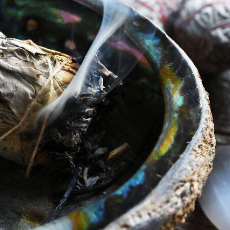 A close up image of a smoldering white sage smudge bundle and healing crystal on a dark wooden table.